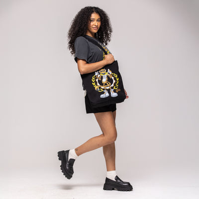 Black tote bag with cartoon Chester Cheetah crest design in gold and white, held by model in black outfit
